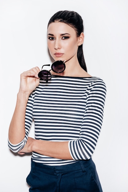 Keeping it cool. beautiful young woman taking off her\
sunglasses and looking at camera while standing against white\
background