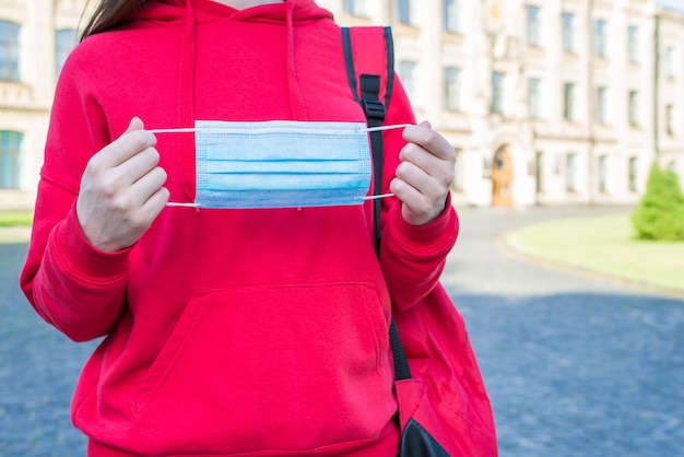 Keeping social distance concept. Cropped close up view photo of young student in casual red jumper holding wearing medical mask