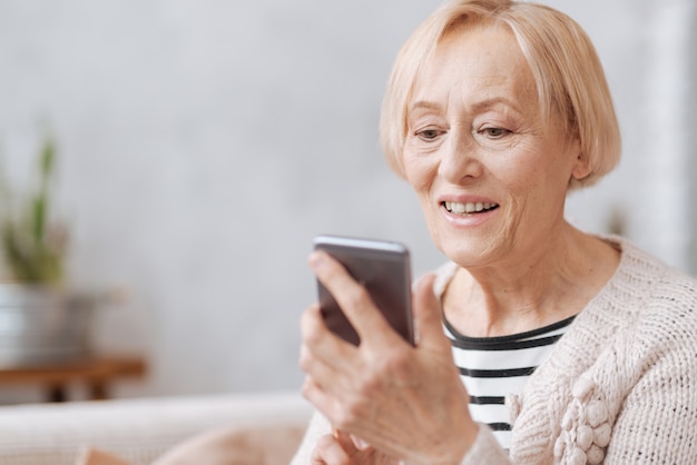 Keeping up with time. Smiling friendly aged lady holding her smartphone in her hands and typing something while spending time at home