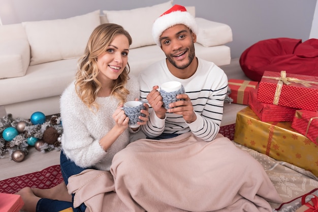 Keeping warmth. Cheerful good looking young couple sitting on the floor and covering their legs with a blanket while enjoying their tea