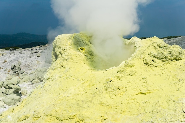 Kegel van zwavelafzettingen rond een fumarole in een solfatarisch veld verlicht door de zon tegen een stormachtig landschap in de verte