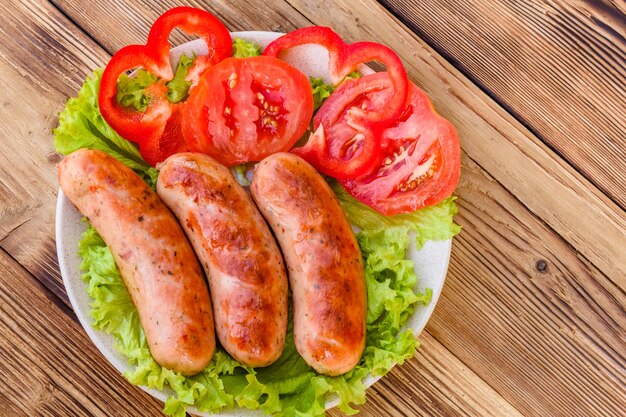 Keramische plaat met gegrilde worstjes, gesneden tomaten en slabladeren op rustieke houten tafel. Bovenaanzicht