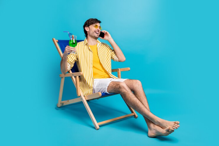 Kerel ik paradijs. Volledige lengte foto man ontspannen rust resort zitten chaise-lounge drinken cocktail bellen vriend vertellen weekend dragen wit geel shirt korte broek blootsvoets geïsoleerd blauwe kleur achtergrond