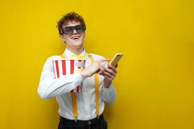 Kerel in feestelijke outfit in 3D-bril kijkt film met popcorn en gebruikt smartphone