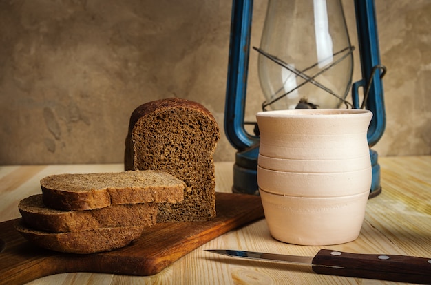 Kerosene lantern cutting board clay pot rye bread and a knife on a wooden table