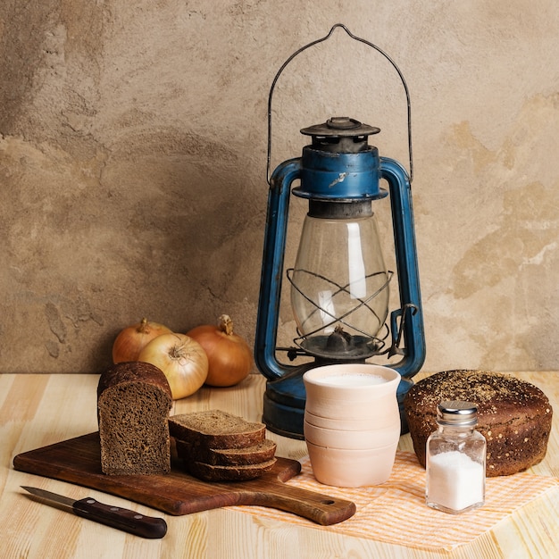 Керосиновый фонарь, разделочная доска, глиняный горшок с молоком, ржаной хлеб, соль и соль и лук на деревянном столе