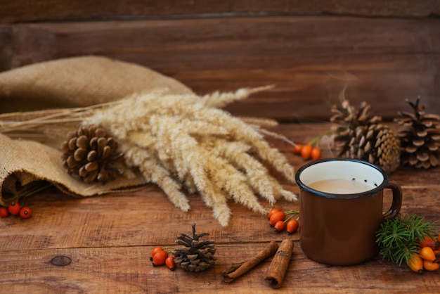 Kerst achtergrond in rustieke stijl. Een metalen mok met hete melkthee staat op een tafelkleed, op een houten ondergrond tussen dennenappels, sparren takken, aartjes van tarwe en rozenbottels. Ruimte kopiëren, plat leggen