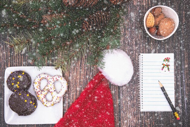 Foto kerst achtergrond met een leeg stuk papier een pen koekjes santas hoed en sneeuwvlokken