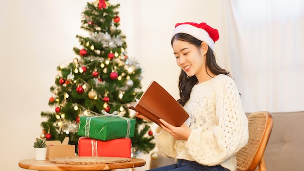 Kerst concept Jonge Aziatische vrouw zittend op de stoel in de buurt van de kerstboom en het lezen van een boek