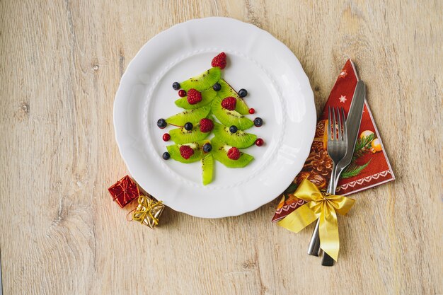 Kerst eten voor kinderen. witte schotel met kerstboom gemaakt van kiwi, frambozen, bosbes en bosbes op houten tafel. Hoge kwaliteit foto