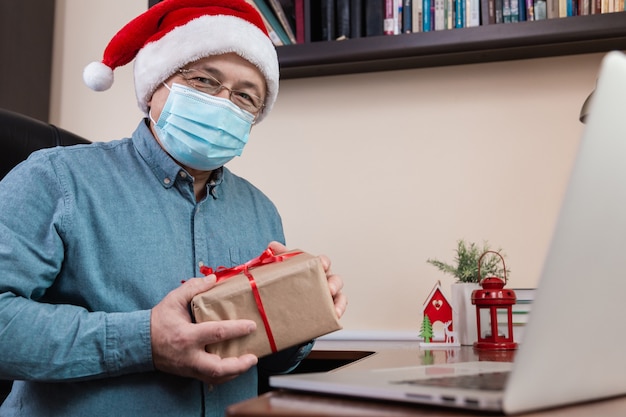 Kerst online felicitatie. Senior man in kerstman hoed en gezichtsmasker Geeft een geschenk en praat met behulp van laptop voor video-oproep vrienden en kinderen. Kerst tijdens coronavirus.
