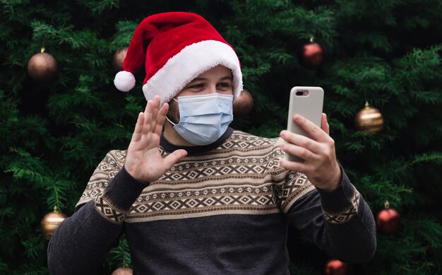 Kerst online gefeliciteerd. Portret van man met kerstmuts en medisch masker praten aan de telefoon met vrienden en ouders. kerstboom bokeh op achtergrond