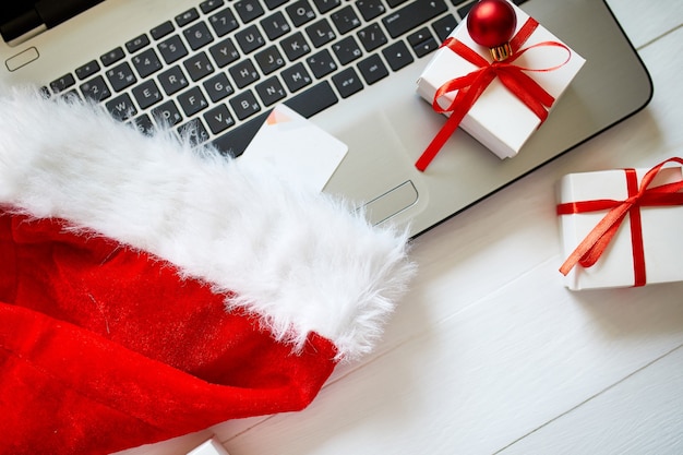 Kerst online winkelen thuis, laptop, creditcard, kerstmuts en geschenkdoos op witte houten tafel, bovenaanzicht