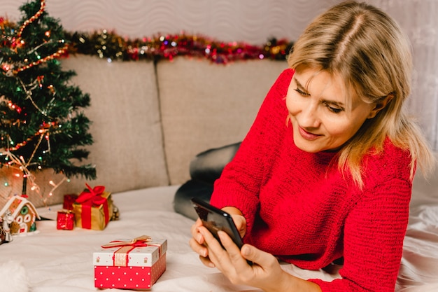 Kerst online winkelen. Vrouwelijke koper maakt bestelling op mobiele telefoon. Vrouw koopt cadeautjes, bereidt zich voor op Kerstmis, geschenkdoos in de hand. Wintervakantie verkoop.