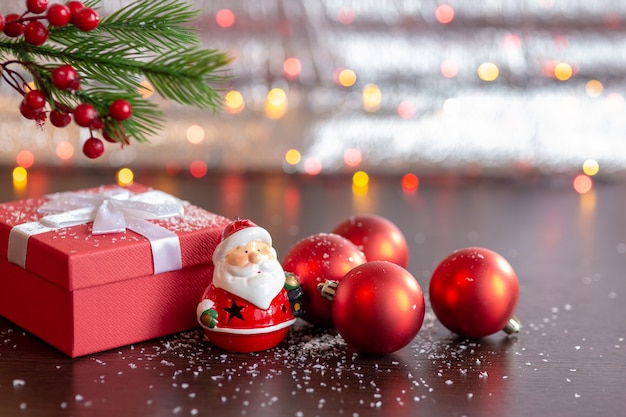 Kerst rode geschenkdoos, een beeldje van de kerstman, rode kerstballen, onder een vuren tak. op een donkere tafel met sneeuw en bokeh van slingers op een lichte bokeh achtergrond.