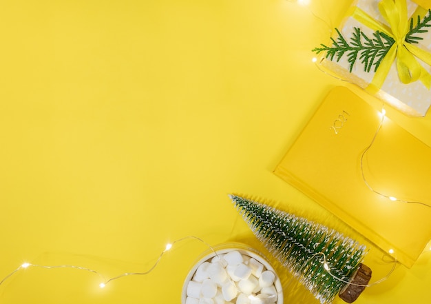 Kerst werkruimte concept in gele kleur. Kader van notitieblok-organizer op 2021, kleine kerstboom, doe-het-zelf geschenkdoos, kopje cacao met marshmallows, kerstverlichting. Planning. Bovenaanzicht, plat gelegd. Kopieer ruimte.