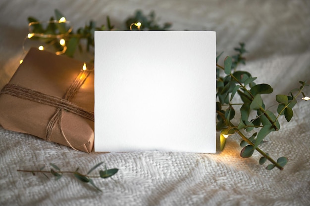 Kerst winter lege 5x5 vierkante kaart mockup met cadeau, eucaliptus bladeren en lichte slinger op een gebreide beige achtergrond, element voor uitnodiging, dank u o wenskaart. Kerst gezellige achtergrond