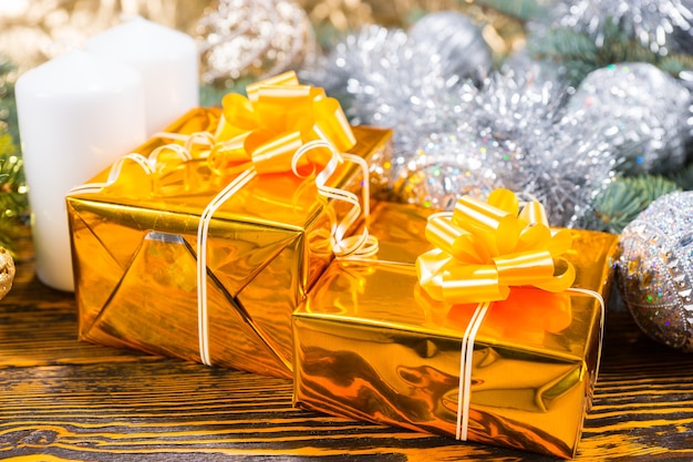 Foto kerstarrangement met twee geschenkdozen verpakt in glanzend goudkleurig papier, witte kaarsen en dennentak versierd met zilveren kerstballen en klatergoud, op een bruin houten tafel