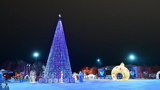 Kerstboom versierd met flikkerende gloeilampen slingers kerstballen Santa Claus Snow Maiden op kerst besneeuwd plein