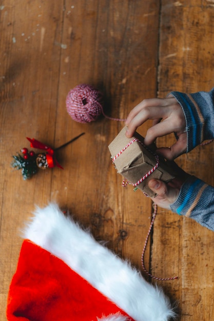 Kerstcadeau verpakking. Handen wikkelen een kleine geschenkdoos in ambachtelijk papier en een rood-wit touwtje op de rustieke houten tafel met een kerstmuts. Sfeervol beeld, Scandinavische nordic style