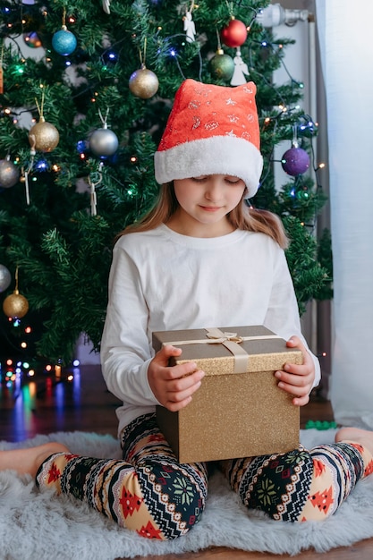 Kerstcadeaus voor kinderen. Een schattig Kaukasisch meisje met een nieuwjaarshoed opent een doos met een cadeau onder een nieuwjaarsboom. Verticale oriëntatie.