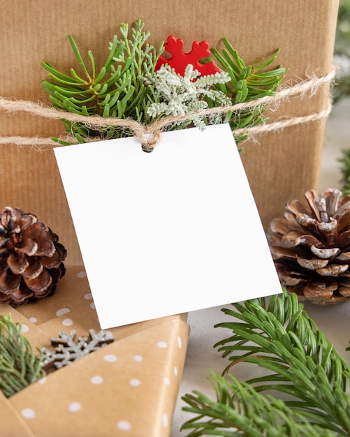 Kerstcadeautje met vierkante papieren cadeaulabel op een witte tafel met dennentakken en decoraties van dichtbij. Rustieke wintercompositie met lege Gift tag Mockup, kopieer ruimte