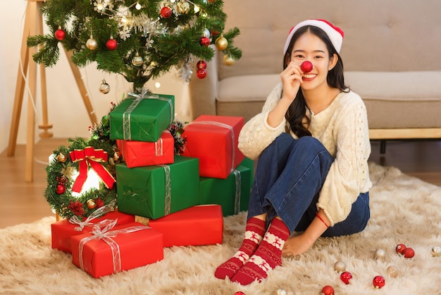 Kerstconcept jonge aziatische vrouw in kerstmuts die glimlacht en rode bal vasthoudt om de neus te bedekken