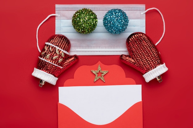 Kerstdecoratie bestaande uit een rode kerst envelop met een wit blanco briefhoofd en twee multi gekleurde kerstballen geplaatst op een medisch masker en de achtergrond is rood