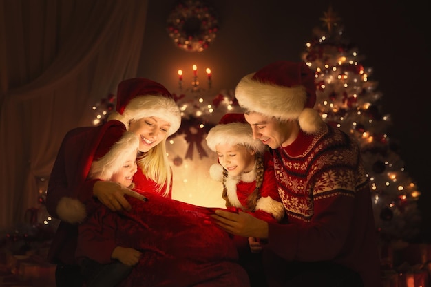 Kerstfamilie die Santa Bag met geschenken opent. Gelukkige ouders en kinderen zitten voor versierde Xmas Fir Tree en kijken naar Magic Light. Wintervakantieavond