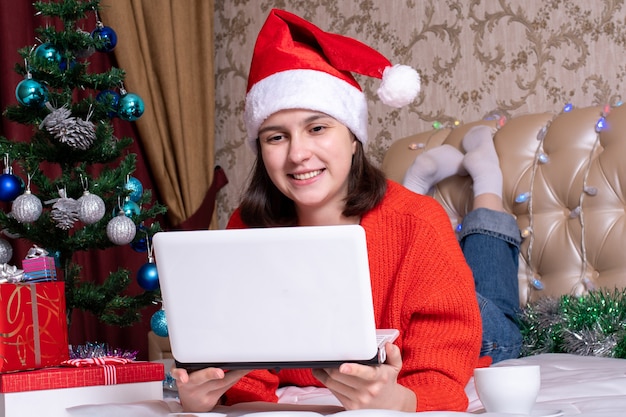 Kerstgroeten op internet, onderwijs, online winkelen. Meisje in een kerstmuts met een laptop thuis, versierde kerstboom, geschenken. Vrolijk kerst- en nieuwjaarsconcept