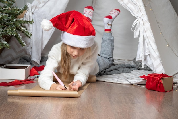 Kersthelperkind dat een brief schrijft aan de kerstman in een rood hoed glimlachend meisje dat een verlanglijstje of een brief aan de kerstman thuis maakt