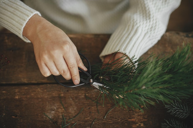 Kerstkrans maken Vrouw in gezellige trui die dennentak met een schaar op rustieke achtergrond snijdt