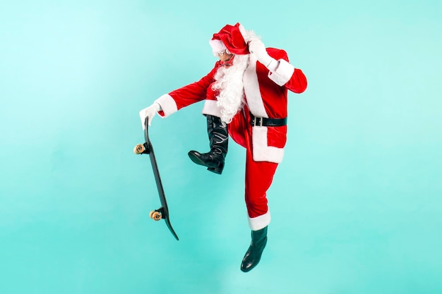 Kerstman in zonnebril en pak springt en vliegt met skateboard op blauwe geïsoleerde achtergrond