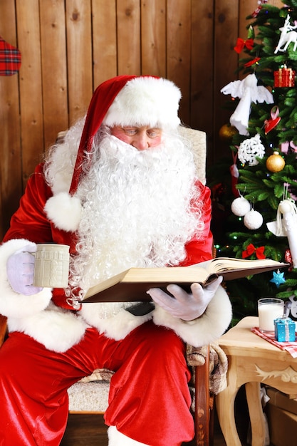 Kerstman met boek zittend in comfortabele schommelstoel in de buurt van Kerstmis thuis at