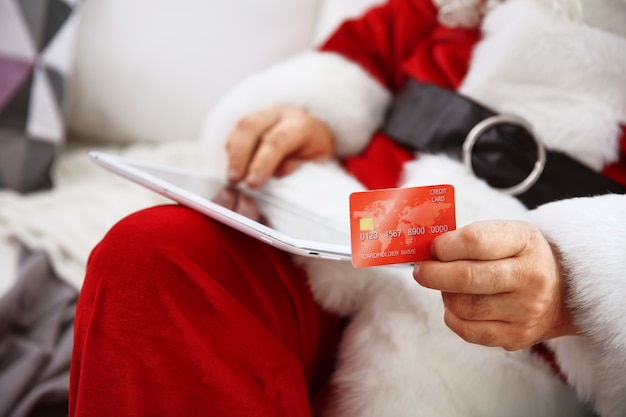 Kerstman met creditcard en tablet op de bank thuis