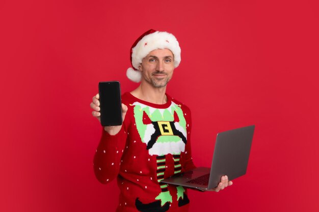 Kerstman met telefoonkaart en laptop Studioportret van man draagt kerstmuts en wintertrui Man met kerstmuts op geïsoleerde rode achtergrond