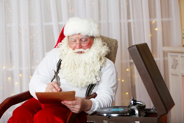 Kerstman zit met een lijst met kinderwensen in een comfortabele stoel bij de open haard thuis