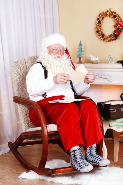 Kerstman zit met kinderbrieven in een comfortabele stoel bij de open haard thuis