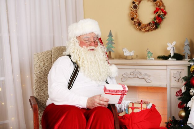 Kerstman zit met kinderen presenteert in een comfortabele stoel bij de open haard thuis