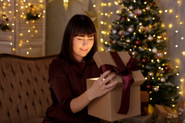 Kerstmis en magisch concept - portret van een jonge vrouw die een magische geschenkdoos opent in een versierde kamer met kerstboom en led-verlichting voor de feestdagen