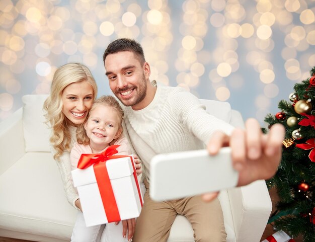 kerstmis, feestdagen, technologie en mensen concept - gelukkig gezin zitten op de bank en het nemen van een selfie foto met smartphone over lichten achtergrond