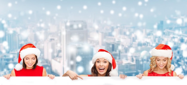 Kerstmis, Kerstmis, mensen, reclame en verkoopconcept - gelukkige vrouwen in de hoed van de Kerstmanhelper met leeg wit bord over de achtergrond van de besneeuwde stad