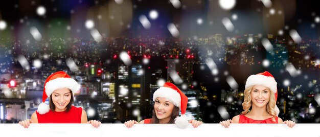 Foto kerstmis, kerstmis, mensen, reclame en verkoopconcept - gelukkige vrouwen in de hoed van de kerstmanhelper met leeg wit bord over de achtergrond van de besneeuwde stad