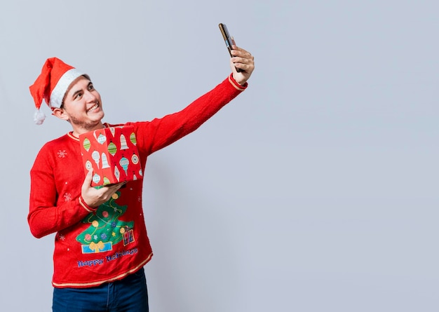 Kerstmis knappe man die een selfie neemt met een cadeau geïsoleerd Gelukkige kerel die een kerst selfie geïsoleerd neemt Lachende jonge man in kerstmuts die een cadeau vasthoudt en een selfie neemt geïsoleerd