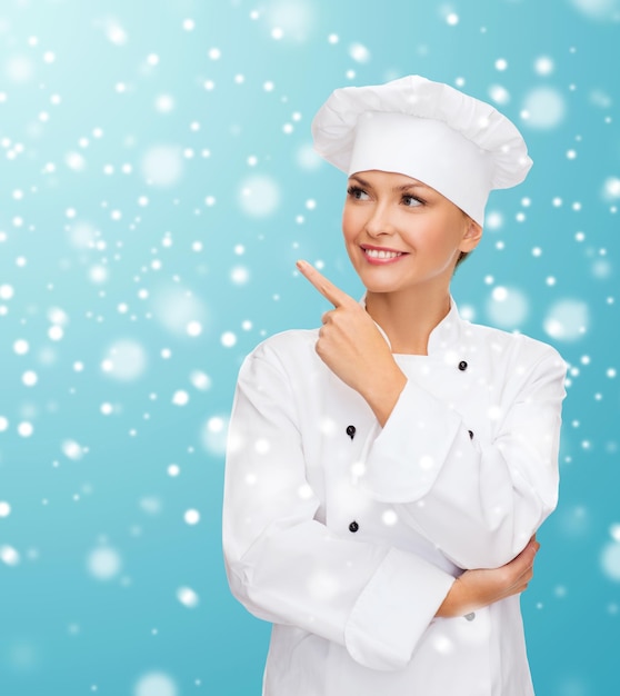 Kerstmis, koken, vakantie en mensen concept - glimlachende vrouwelijke chef-kok, kok of bakker wijzende vinger omhoog over blauwe besneeuwde achtergrond