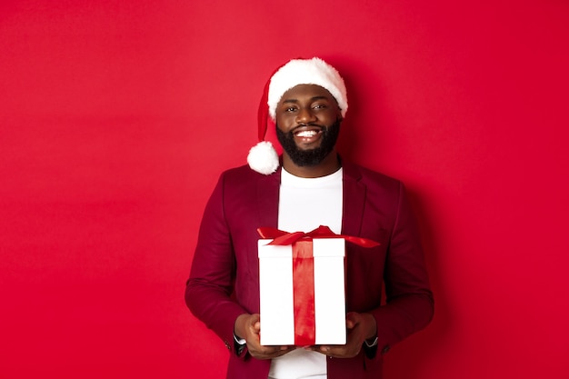 Kerstmis, Nieuwjaar en winkelconcept. Gelukkig bebaarde Afro-Amerikaanse man met kerstcadeau, glimlachend in de camera, staande in kerstmuts tegen rode achtergrond