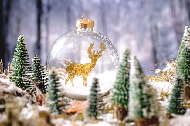 Kerstmis of Nieuwjaar wenskaart. Glazen transparante bal gouden hert binnen met decoratieve kerstbomen rond op besneeuwd mos met winterbos op de achtergrond. Kerstvakantie sfeer