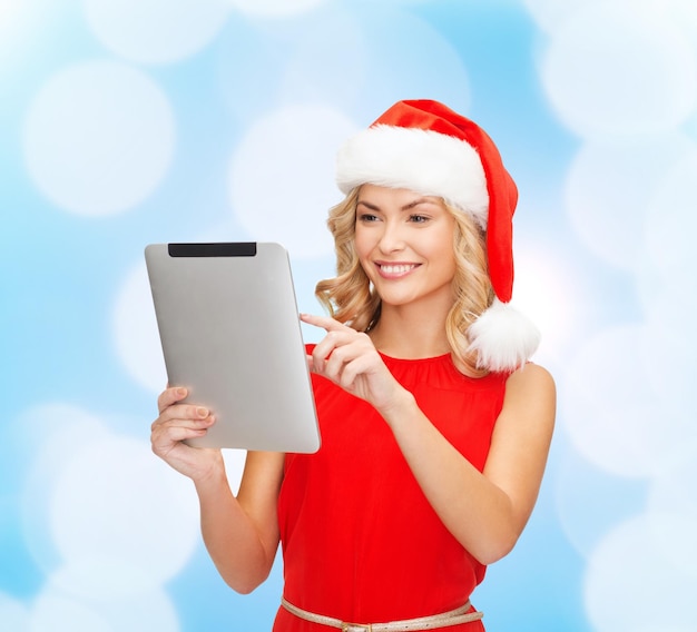 Kerstmis, technologie, heden en mensenconcept - glimlachende vrouw in de hoed van de kerstman met de computer van tabletpc over blauwe lichtenachtergrond
