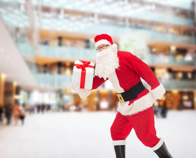 Kerstmis, vakantie en mensen concept - man in kostuum van de kerstman loopt met geschenkdoos over winkelcentrum achtergrond