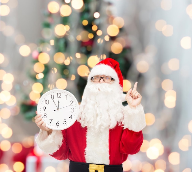 Kerstmis, vakantie en mensen concept - man in kostuum van de kerstman met klok met twaalf wijzende vinger omhoog over boom lichten achtergrond